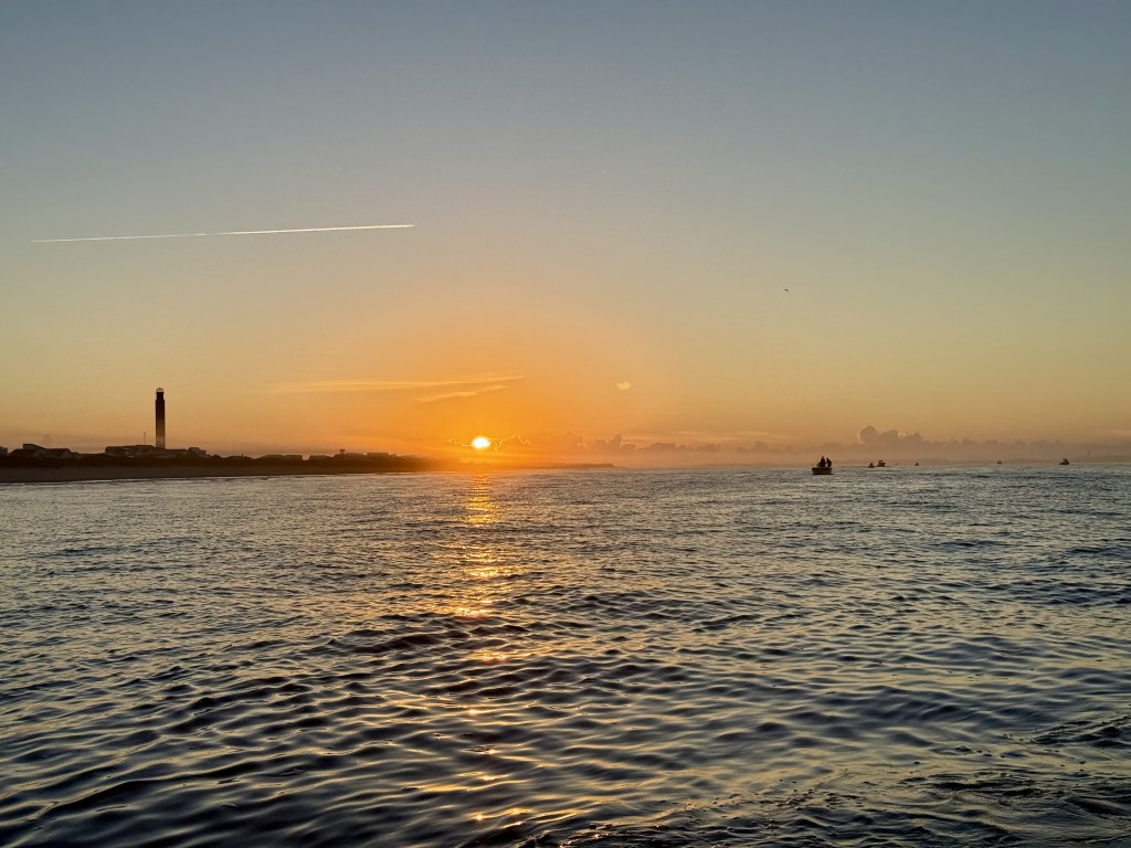 Sunset over the ocean with rippling water and a distant lighthouse silhouette.